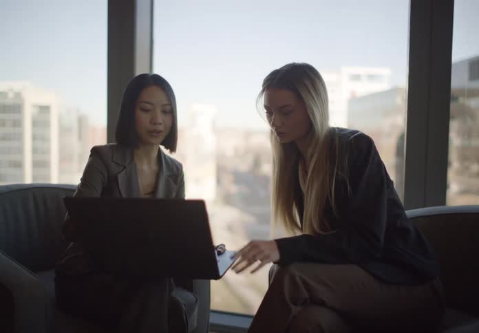 Two diverse businesswomen collaborate on a project using a laptop in a modern, sunlit office lounge with a city view