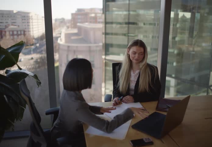 Two diverse businesswomen collaborate on a project in a modern city office, using a laptop and reviewing documents