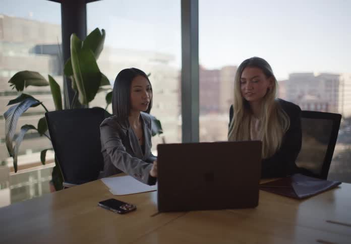 Two diverse professional women collaborate on a laptop during a meeting in a modern office with a city view