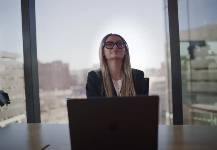 A focused businesswoman works on her laptop, then pauses to think before leaning back with a satisfied expression in her modern city office