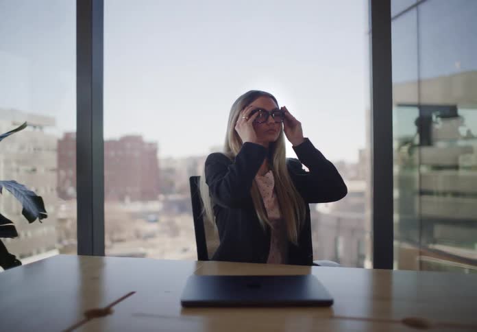 A young professional woman closes her laptop and puts on her glasses while contemplating in a modern city conference room