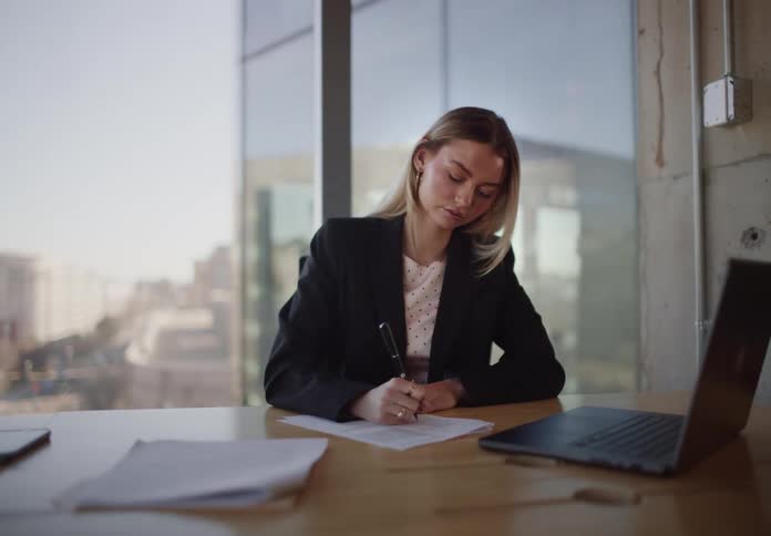 A tired businesswoman works on paperwork at her desk in a modern office with a city view