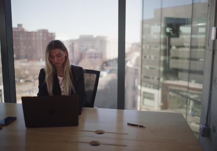 A slow zoom-out shot of a focused young businesswoman working on her laptop in a modern high-rise office overlooking the city