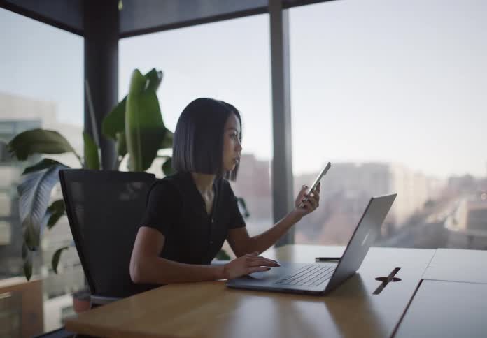 High-angle shot pulls back from a businesswoman analyzing charts on her phone and laptop in a modern city office