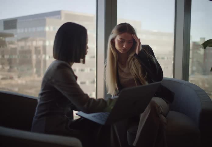 Two diverse female colleagues collaborate on a project using a laptop in a modern city office, one explaining while the other listens intently with a serious expression