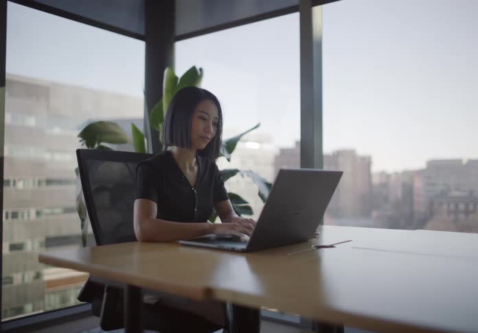 Stylish Asian businesswoman participates in a video call then works on her laptop in a modern corner office with a city view