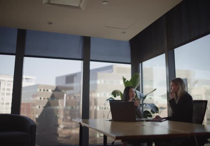 Two professional businesswomen collaborate during a meeting in a modern corner office with large windows overlooking the city