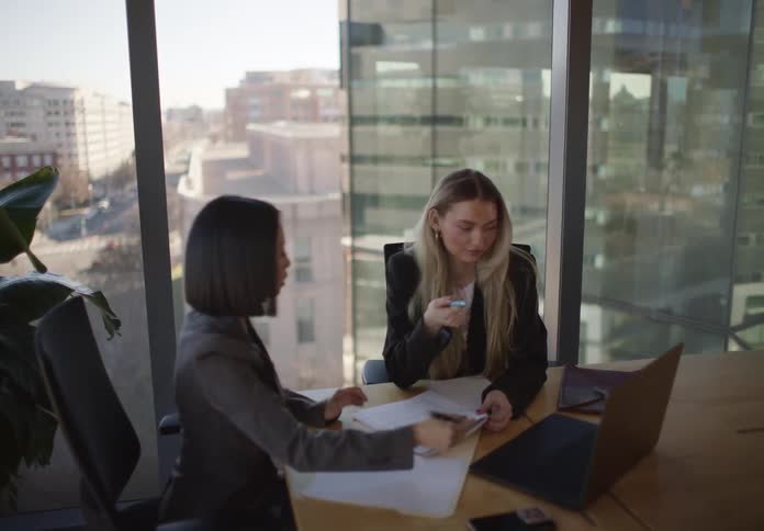 Two diverse female colleagues collaborate in a modern office with a city view, discussing work on a laptop and taking notes