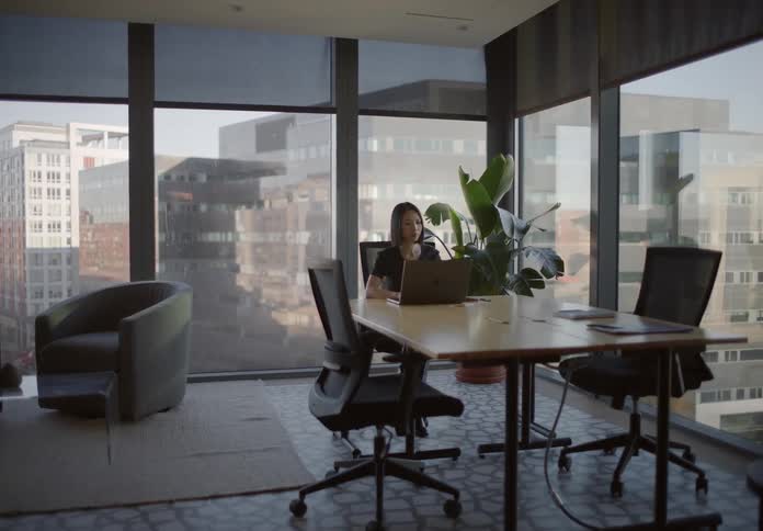 A tracking shot zooms in on a professional woman working on a laptop in a modern, sunlit office with a city view