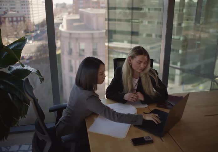 Two professional women collaborate on a project in a bright, modern office with a view of the city, reviewing documents and working together on a laptop