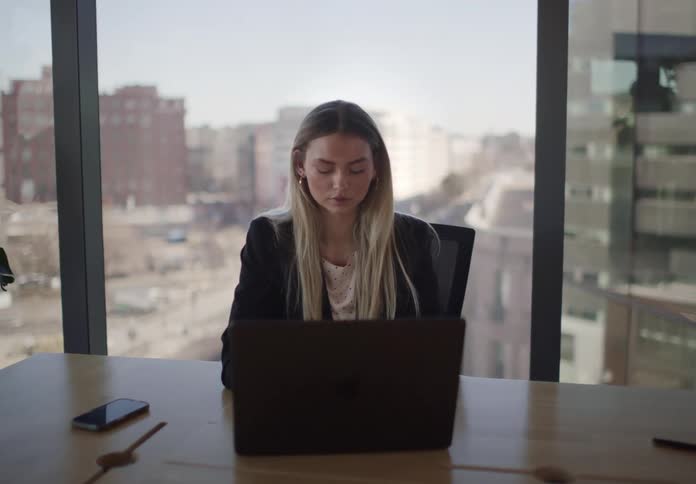 A slow zoom-in shot shows a focused young businesswoman working on her laptop at a desk in a high-rise office with a city view