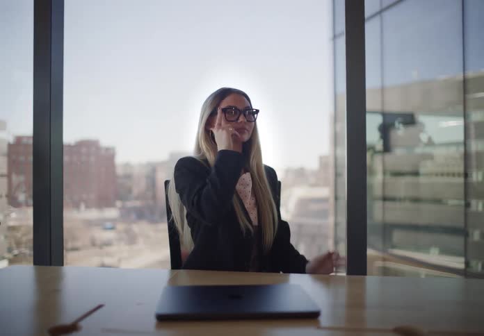 A confident businesswoman closes her laptop, puts on her glasses, and reflects while sitting in a modern high-rise office