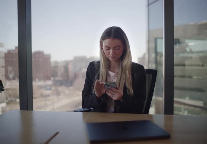 A slow zoom on a young businesswoman scrolling on her smartphone while sitting in a modern high-rise office with a city view