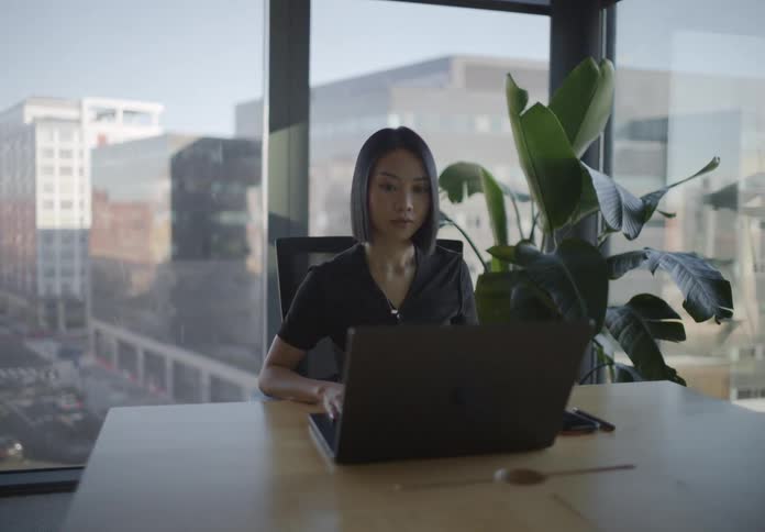A stylish young professional woman enters a modern city office, sits at a conference table with a view, and begins working on her laptop