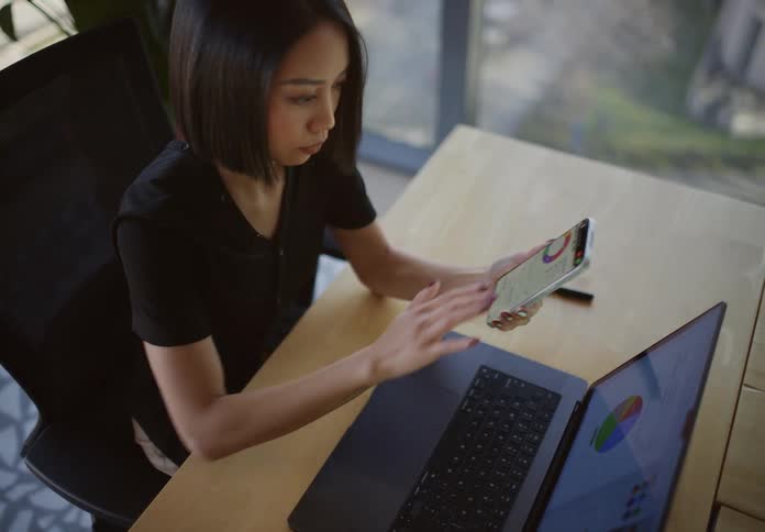A young professional woman analyzes business data and charts on her laptop and smartphone while working at her desk