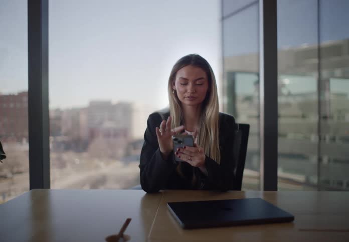 A young businesswoman closes her laptop after finishing work and scrolls on her smartphone in a modern, sunlit office