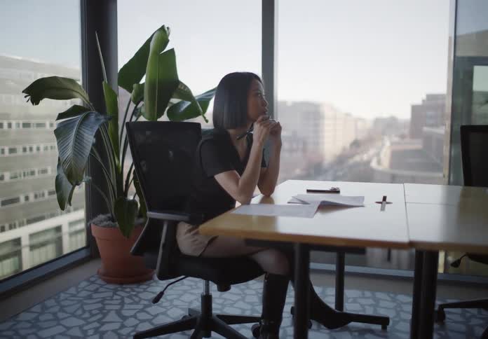 A stylish young Asian woman sits at a desk in a modern city office, thoughtfully looks out the window, and then begins to write on a document