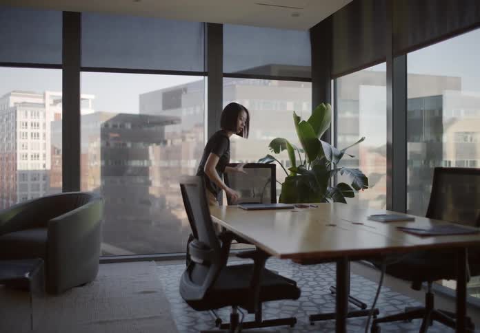 A stylish Asian businesswoman walks in slow motion through a modern office, sits at a conference table with a city view, and begins working on her laptop