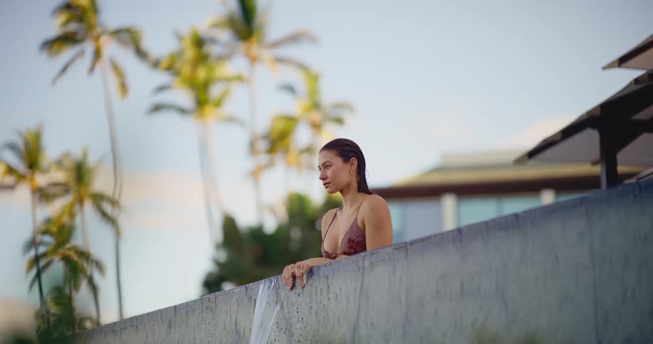 A serene woman in a bikini relaxes at the edge of an infinity pool in slow motion with palm trees in the background