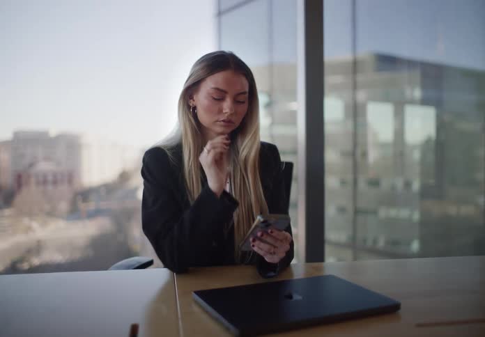 A focused businesswoman closes her laptop to scroll on her smartphone while sitting in a modern office conference room with a city view