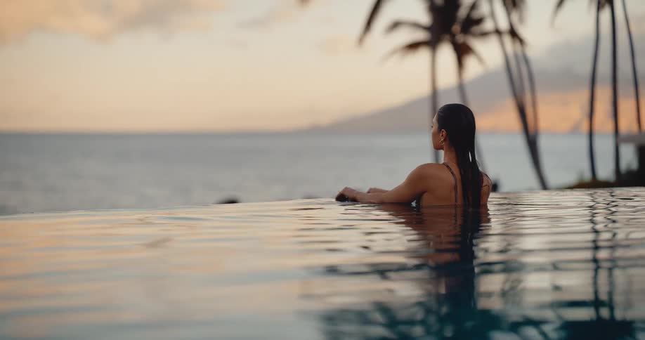 A beautiful woman relaxes in a luxury resort infinity pool at sunset, looking out at the tranquil ocean and palm trees