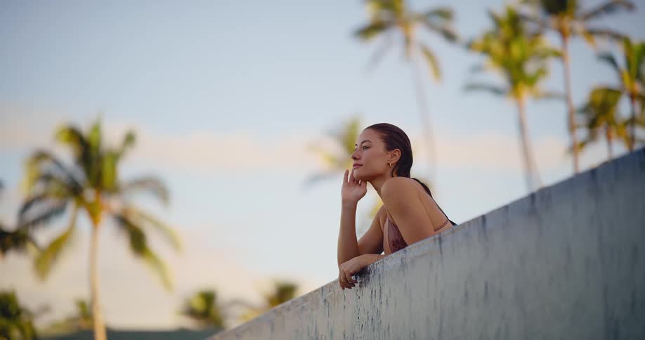 Contemplative young woman in a bikini rests on the edge of a rooftop infinity pool, enjoying the tranquil tropical scenery