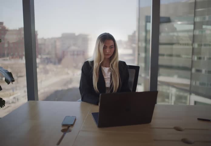 A thoughtful young businesswoman works on her laptop in a sunlit modern office overlooking the city