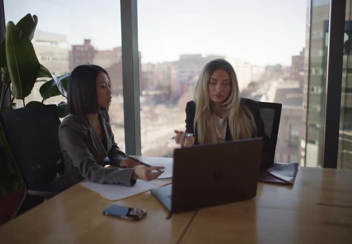 Slow zoom into two diverse businesswomen collaborating on a project in a modern office overlooking the city