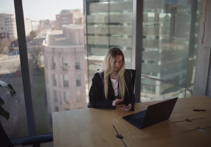 A young businesswoman works on her laptop and checks her phone in a modern city office with large windows