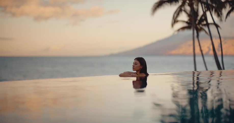 A serene woman relaxes in a luxury infinity pool, enjoying the tranquil tropical sunset view over the ocean with palm trees in the background