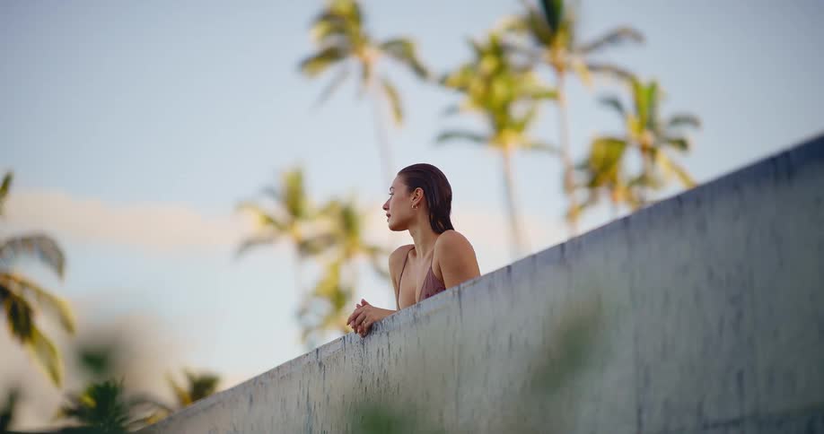 A serene woman in a bikini enjoys the tropical view from the edge of an infinity pool with palm trees in the background