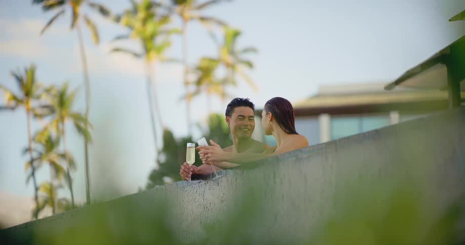 Happy young couple enjoys champagne together while relaxing in a beautiful infinity pool at a luxury tropical resort