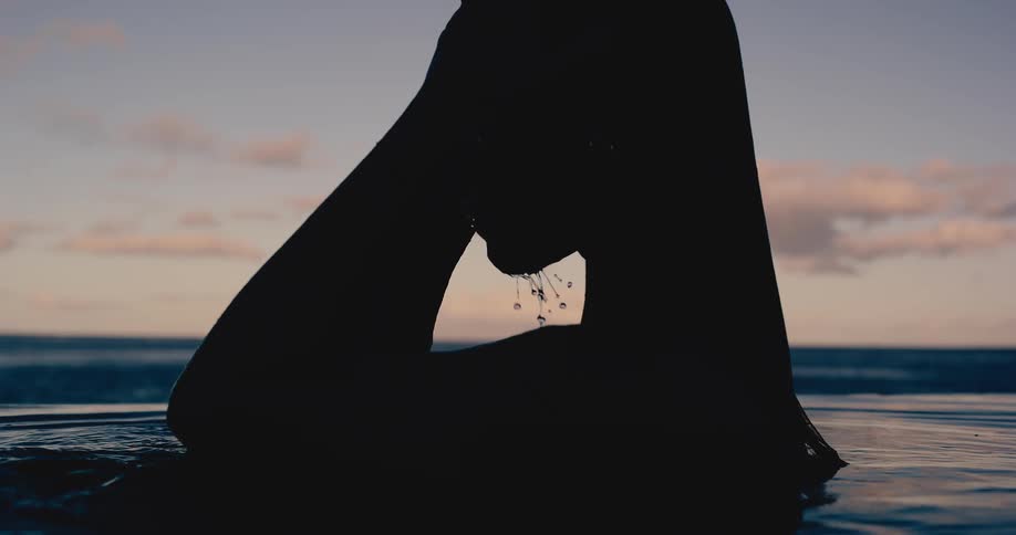 A silhouetted woman slowly emerges from an infinity pool at dusk, running her hands through her wet hair in super slow motion