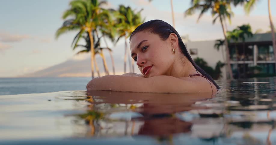 A beautiful young woman with wet hair looks at the camera while relaxing at the edge of a luxurious infinity pool at a tropical resort