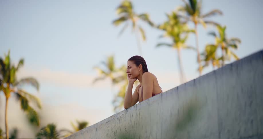 A tranquil woman in a bikini relaxes at the edge of a swimming pool in slow motion with tropical palm trees in the background
