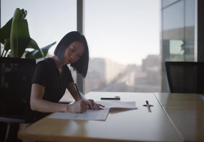 An Asian businesswoman sits alone in a modern conference room, signing documents with a city view