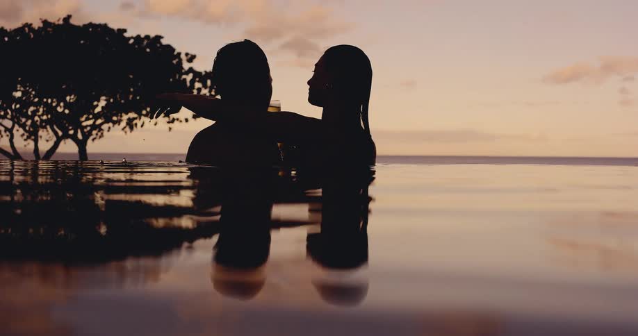 A silhouetted couple romantically enjoys champagne in a luxurious infinity pool as the sun sets over the ocean