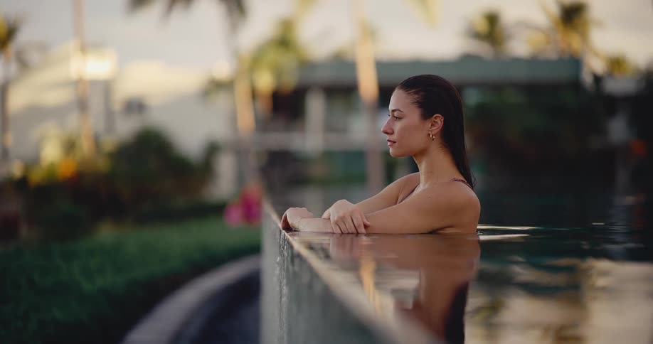 A serene woman with wet hair relaxes at the edge of a resort infinity pool at sunset
