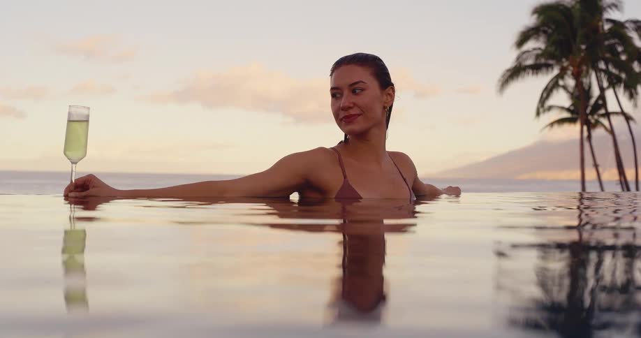 A beautiful young woman in a bikini relaxes with a glass of champagne in a luxurious infinity pool overlooking the ocean at sunset