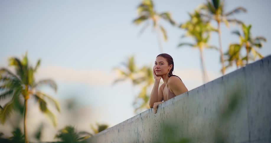 A beautiful young woman relaxes at the edge of a pool, enjoying a peaceful moment at a tropical resort with palm trees in the background