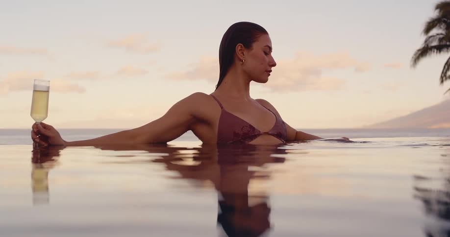 Beautiful woman relaxes in a luxury infinity pool with a glass of champagne, enjoying a tropical sunset over the ocean