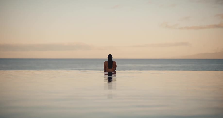 A woman emerges from a tranquil infinity pool, peacefully watching the serene ocean at sunset