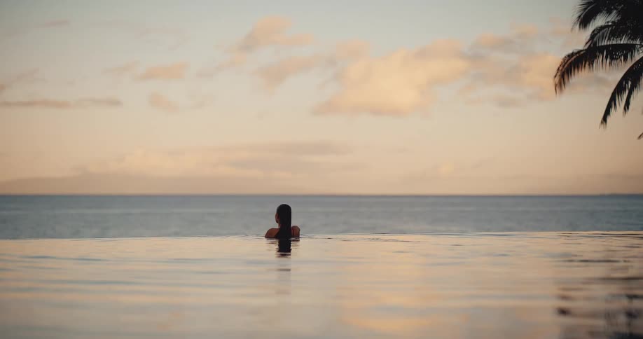 A woman relaxes in a beautiful infinity pool at a tropical resort, enjoying the serene ocean view at sunset