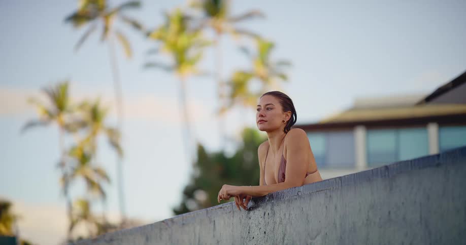 A serene woman in a bikini relaxes at the edge of an infinity pool with palm trees in the background, eventually turning to smile at the camera