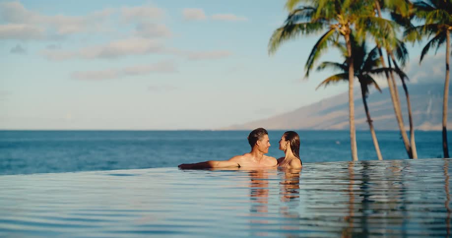 Attractive romantic couple relaxes in a tropical infinity pool at sunset, kissing and smiling