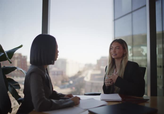 Two professional women, one Asian and one Caucasian, collaborate during a business meeting in a high-rise office overlooking the city