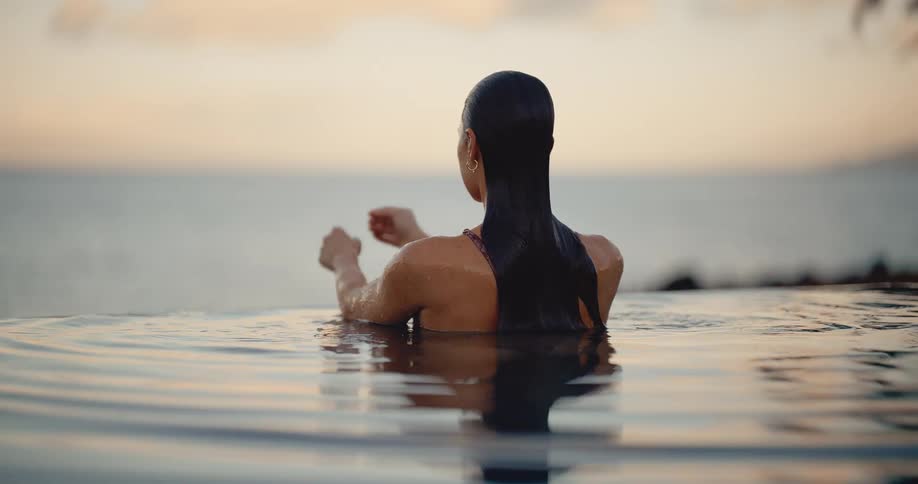 A woman with long wet hair gracefully emerges from an infinity pool in slow motion, enjoying a tranquil sunset over the ocean