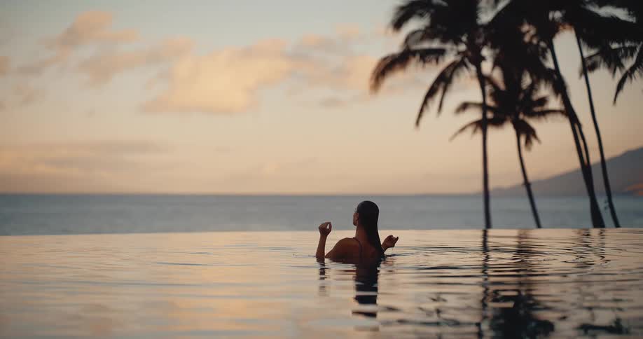 A woman slowly rises from the water of a luxurious infinity pool, relaxing and enjoying the serene tropical sunset over the ocean