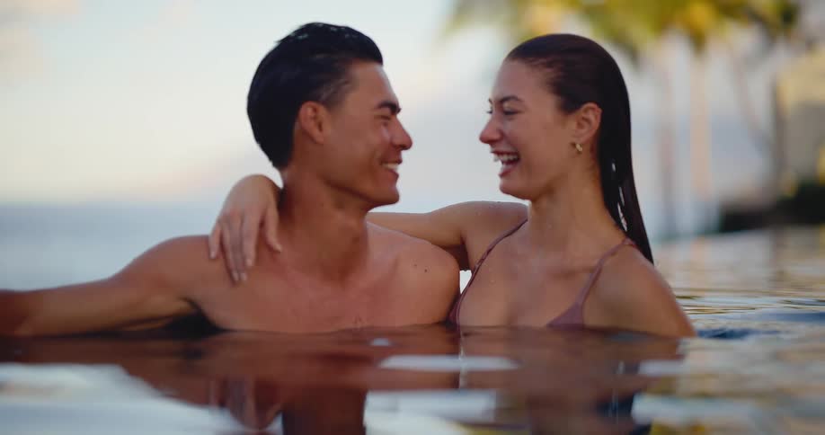 An attractive couple shares an intimate and happy moment, smiling and laughing together in a beautiful infinity pool at a tropical resort