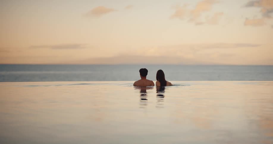 A couple with their backs to the camera peacefully watches the sunset over the ocean from a luxurious infinity pool